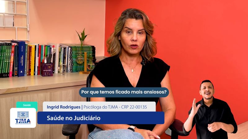 Reprodução de tela de computador com vídeo de programa com apresentadora e intérprete de Libras. A imagem mostra uma mulher branca, de cabelos louros, sentada em uma cadeira, vestindo uma blusa preta e calça jeans clara. O ambiente é interno, com uma parede vermelha ao fundo. À esquerda, há uma estante com vários livros e alguns objetos decorativos, incluindo uma planta. Na parte inferior da imagem, há uma barra de informações que identifica a mulher como Ingrid Rodrigues, psicóloga do TJMA, acompanhada do número de seu registro profissional (CRP 22-00135). 