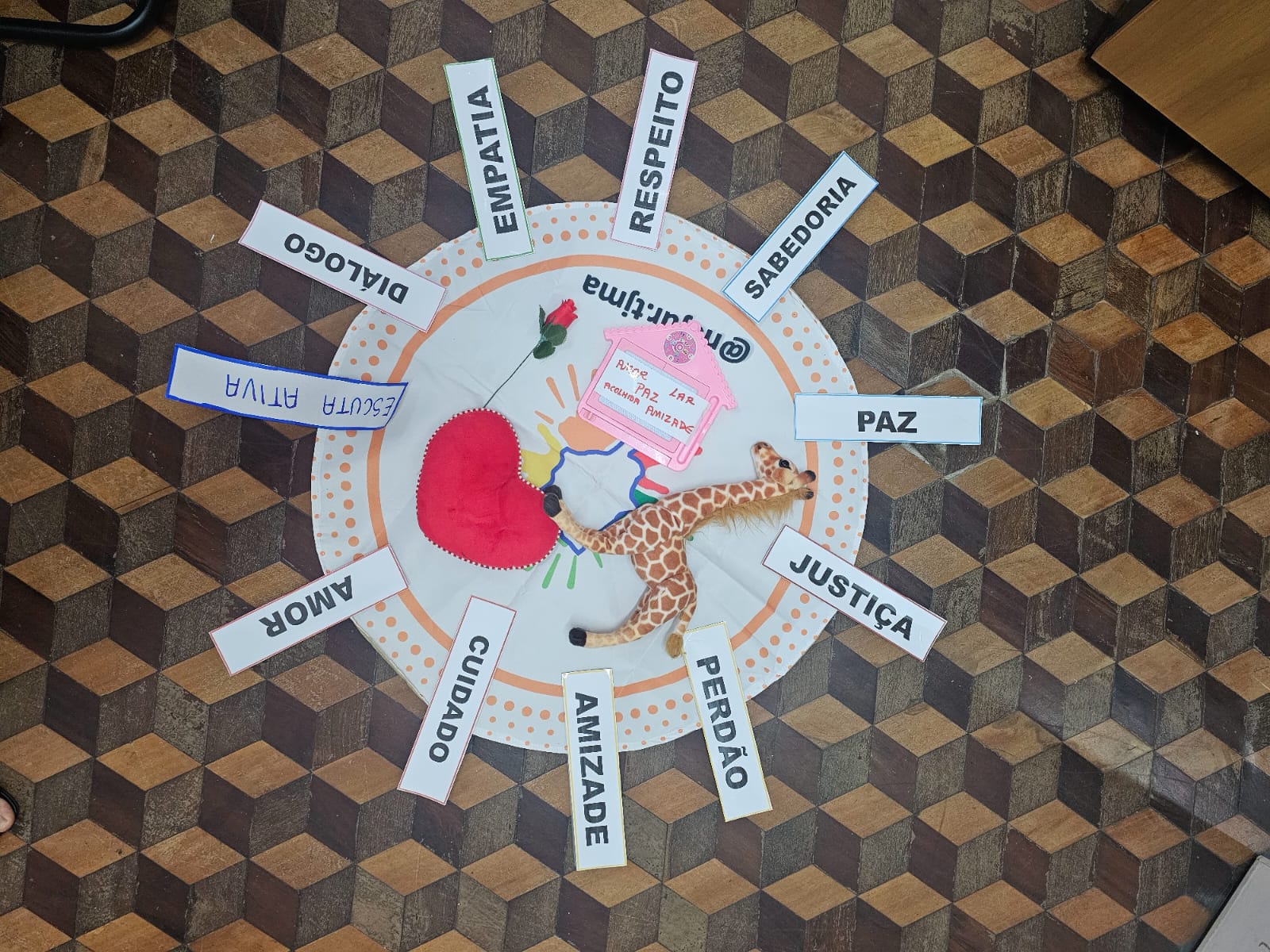 A imagem mostra um círculo no chão, decorado com uma borda laranja pontilhada. Ao redor do círculo, estão distribuídas várias palavras em cartazes retangulares, cada um com um tema positivo: "EMPATIA", "RESPEITO", "SABEDORIA", "PAZ", "JUSTIÇA", "PERDÃO", "AMIZADE", "CUIDADO", "AMOR", "DIÁLOGO" e "ESCUTA ATIVA". No centro do círculo, há um coração vermelho de tecido, uma girafa de brinquedo, uma flor vermelha e uma placa decorativa com as palavras "Respeito", "Paz", "Lar", "Amizade". O chão é de piso de madeira em padrão geométrico.