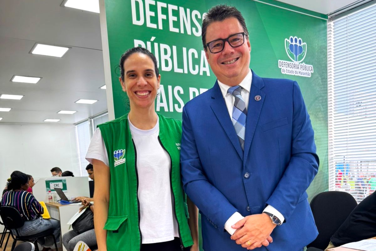 A imagem mostra um grupo de seis pessoas em frente a um banner verde com texto. O banner contém as palavras "DEFESA" e "ESTADO" em destaque, além de "MINISTÉRIO DA JUSTIÇA E SEGURANÇA PÚBLICA" e "Defensoria Pública do Estado do Maranhão." Uma pessoa segura um documento, que parece ser o foco da imagem. Os indivíduos estão vestidos de forma variada, incluindo trajes casuais, um terno e uniformes. O cenário parece ser ao ar livre, com um prédio e janelas ao fundo. A imagem sugere um evento oficial ou institucional, possivelmente relacionado a questões jurídicas ou de defesa pública no estado do Maranhão, Brasil.