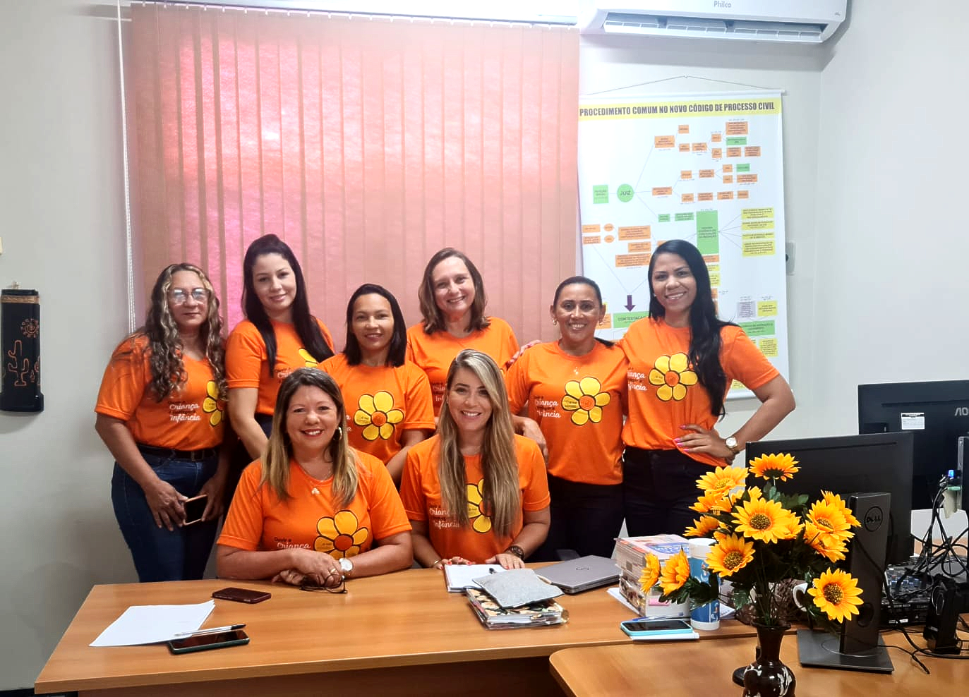 Foto retangular. Mulheres vestidas com camisas laranja da campanha Maio Laranja, com desenho linear de flor, com pétalas amarelas e miolo laranja, em sala fechada.