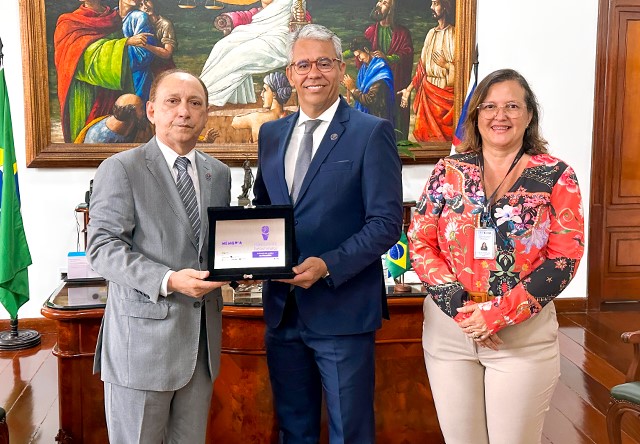 Foto horizontal. No gabinete da Presidência do TJMA, os desembargadores Lourival Serejo e Paulo Velten seguram a placa do o Prêmio Nacional Memória do Poder Judiciário 2023, do Conselho Nacional de Justiça (CNJ). Ao lado deles, a coordenadora de Biblioteca, Cíntia Andrade. Os três estã de frente para a câmera. Lourival Serejo, homem branco, idoso, calvo usa terno cinza claro, gravata em mesmo tom e camisa branca. Paulo Velten, homem branco de meia idade, cabelos grisalhos, óculos de grau, usa terno azul marinho, gravata cinza e camisa branca. Cíntia Andrade, mulher de meia idade, cabelos castanhos, óculos de grau, usa blusa estampada floral e calça bege. A placa tem fundo prata, palavra e letras em liás. O gabinete tem atrás um quadro com personagens antigos, bandeira do bRasil, móvel e piso marrons.