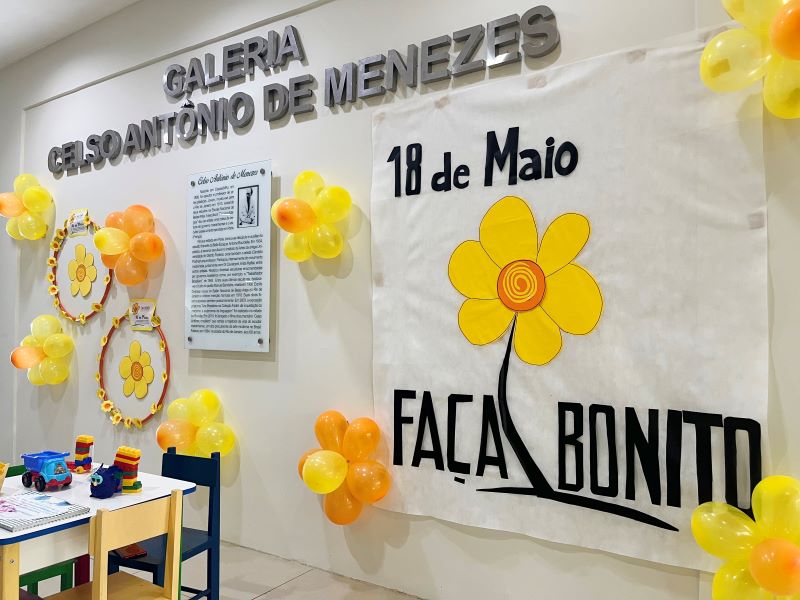Foto colorida de parede com o nome da galeria Celso Antônio de Menezes, com flores amarelas e dizeres 18 de maio, no Fórum de São Luís.