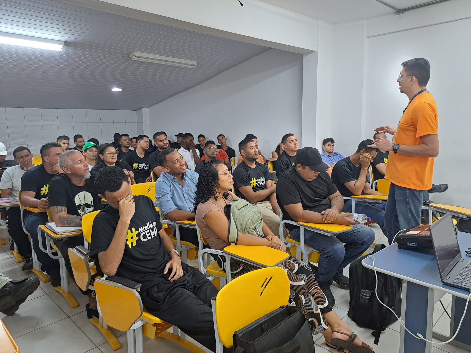 Foto colorida mostra um homem em pé com camisa laranja, Arthur Darub, palestrando para homens e mulheres do Centro de Ensino Médio e Profissionalizante do Maranhão (CEMP).