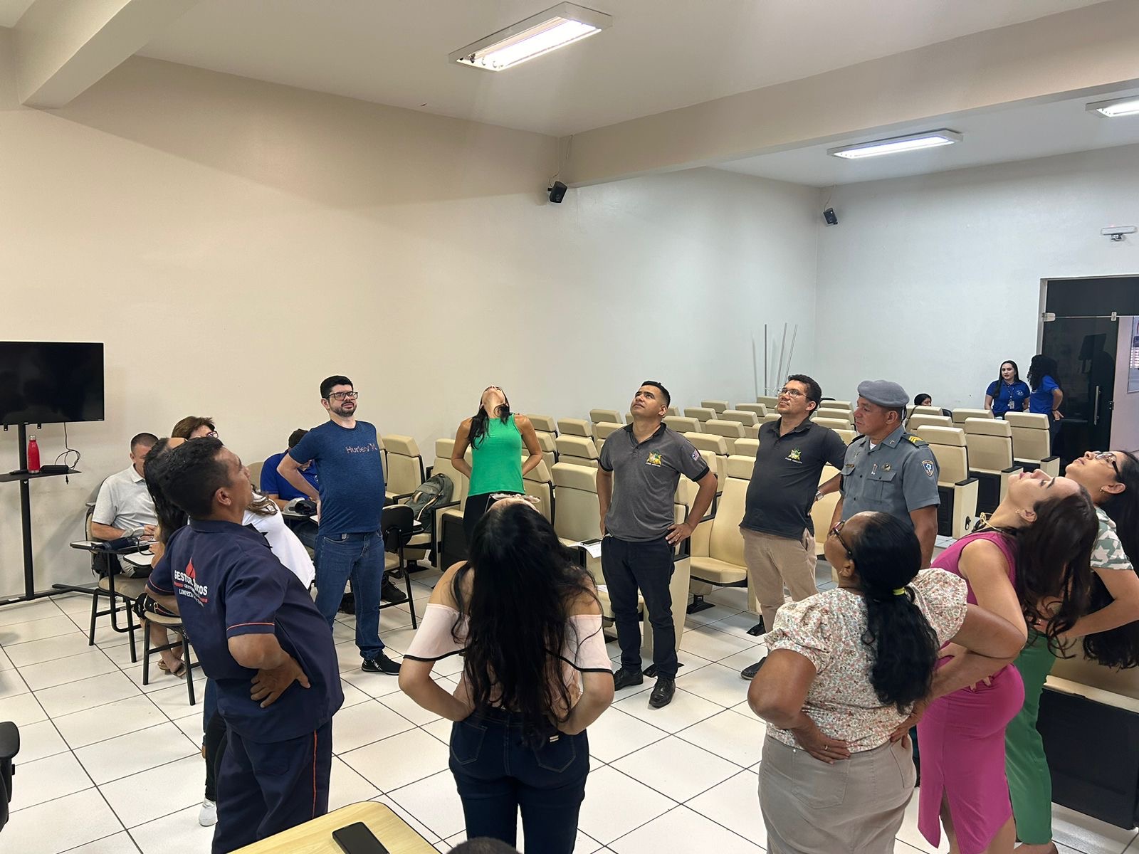 Foto mostra diversas pessoas em pé durante dinâmica de grupo. Todas estão alongando a coluna para trás em um auditório.