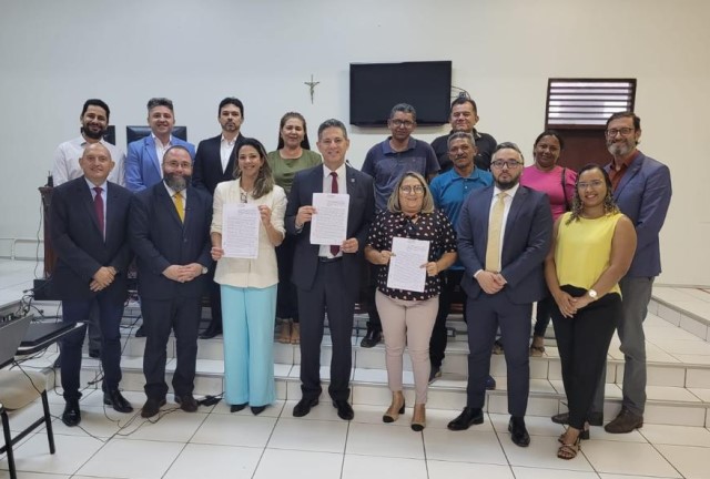 Foto horizontal de acordo fundiário realizado pela Comissão de Conflitos Fundiários do TJMA em Imperatriz. Onze homens e cinco mulheres posam em pé para foto em sala de parede bege e piso branco. Entre as pessoas, três na fila da frente seguram documentos, entre elas o presidente da Comissão de Conflitos Fundiários do TJMA, desembargador Gervásio dos Santos, homem branco de meia idade, que usa terno azul marinho, camisa brabca e gravata vermelha. Na foto, há pessoas branca, pardas e negras. Atrás, um crucifixo na parede e um aparelho de TV.