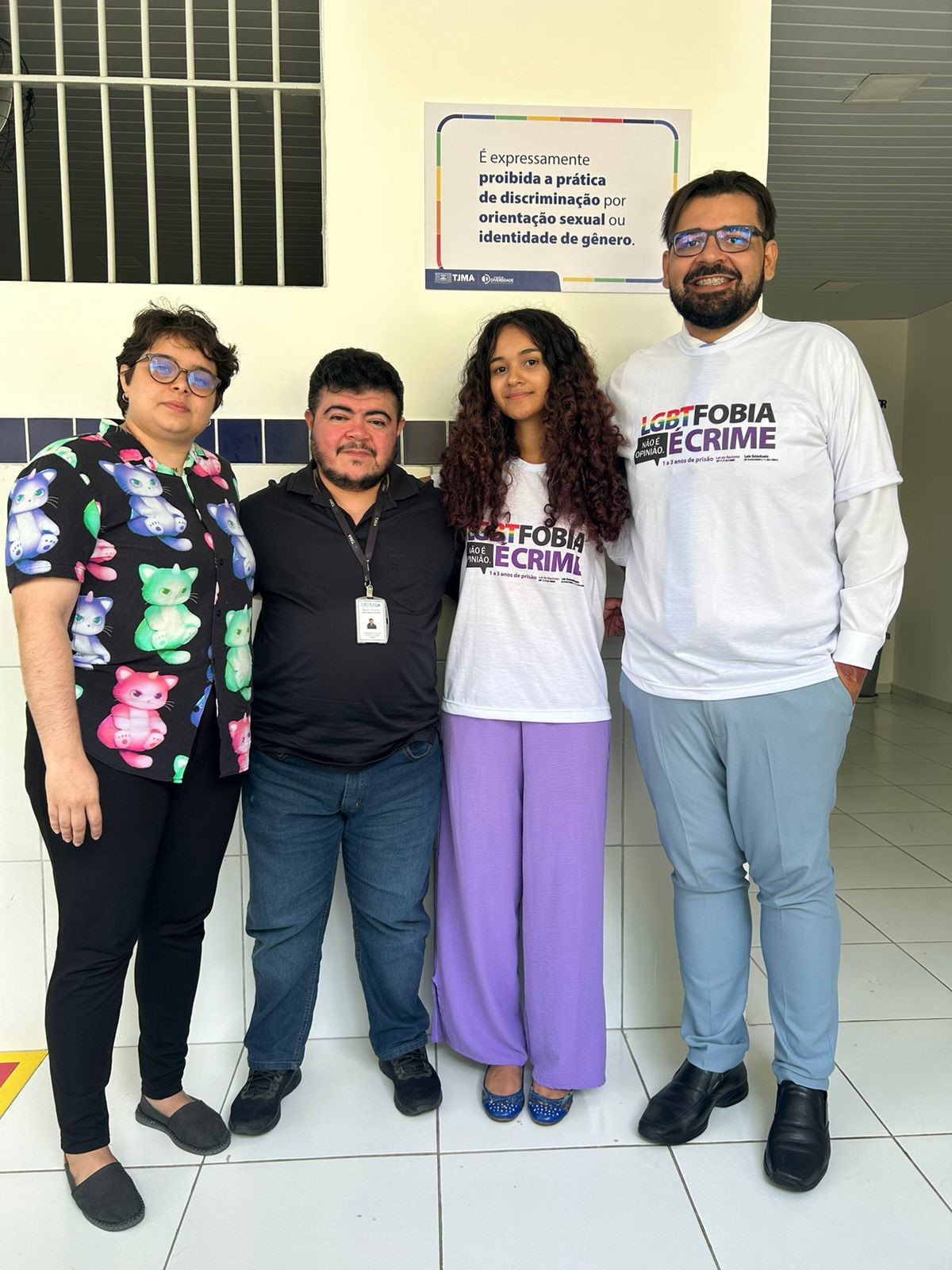 A imagem mostra quatro pessoas lado a lado. Da esquerda para a direita: a primeira pessoa está vestindo uma blusa estampada com personagens de desenho animado, a segunda pessoa está com uma camiseta escura e calças, usando um crachá, a terceira pessoa está com um vestido roxo e segurando um cartaz, e a quarta pessoa está com uma camiseta branca com texto. Atrás delas, há um cartaz na parede que inclui texto relacionado à discriminação e identidade de gênero. Os rostos de todas as pessoas foram ocultados com retângulos marrons para proteger sua privacidade. Essa imagem pode ser relevante ou interessante devido ao seu potencial contexto de abordar ou promover a conscientização sobre questões de discriminação, como sugerido pela mensagem visível no cartaz e na camiseta.