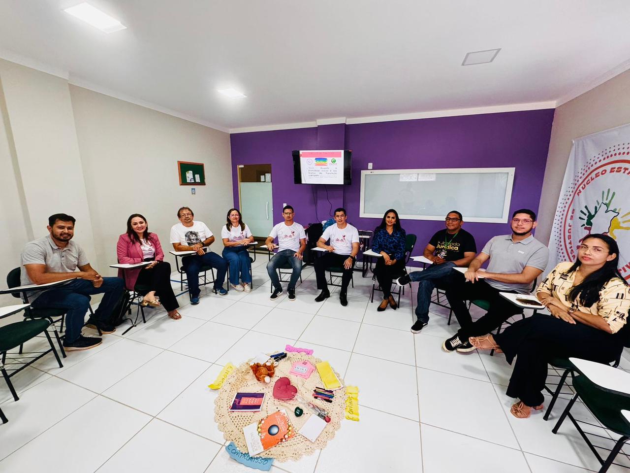 A imagem mostra um grupo de pessoas sentadas em círculo em um ambiente interno, possivelmente durante uma reunião ou workshop. A sala tem paredes roxas e está equipada com um quadro branco e uma tela digital exibindo informações. No centro do círculo, há uma mesa com vários itens, incluindo o que parece ser comida e bebidas. 