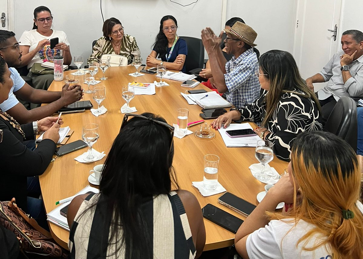 A imagem retrata um grupo de pessoas sentadas ao redor de uma grande mesa de conferência em uma sala de reuniões. Na mesa, há diversos itens, como papéis, cadernos, canetas e garrafas de água, indicando que uma reunião formal ou discussão está ocorrendo.