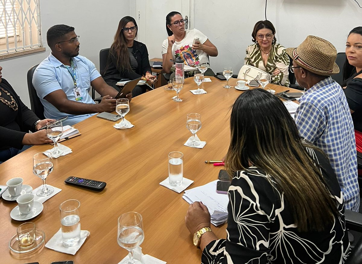 A imagem retrata um grupo de pessoas sentadas ao redor de uma grande mesa de conferência em uma sala de reuniões. Na mesa, há diversos itens, como papéis, cadernos, canetas e garrafas de água, indicando que uma reunião formal ou discussão está ocorrendo.