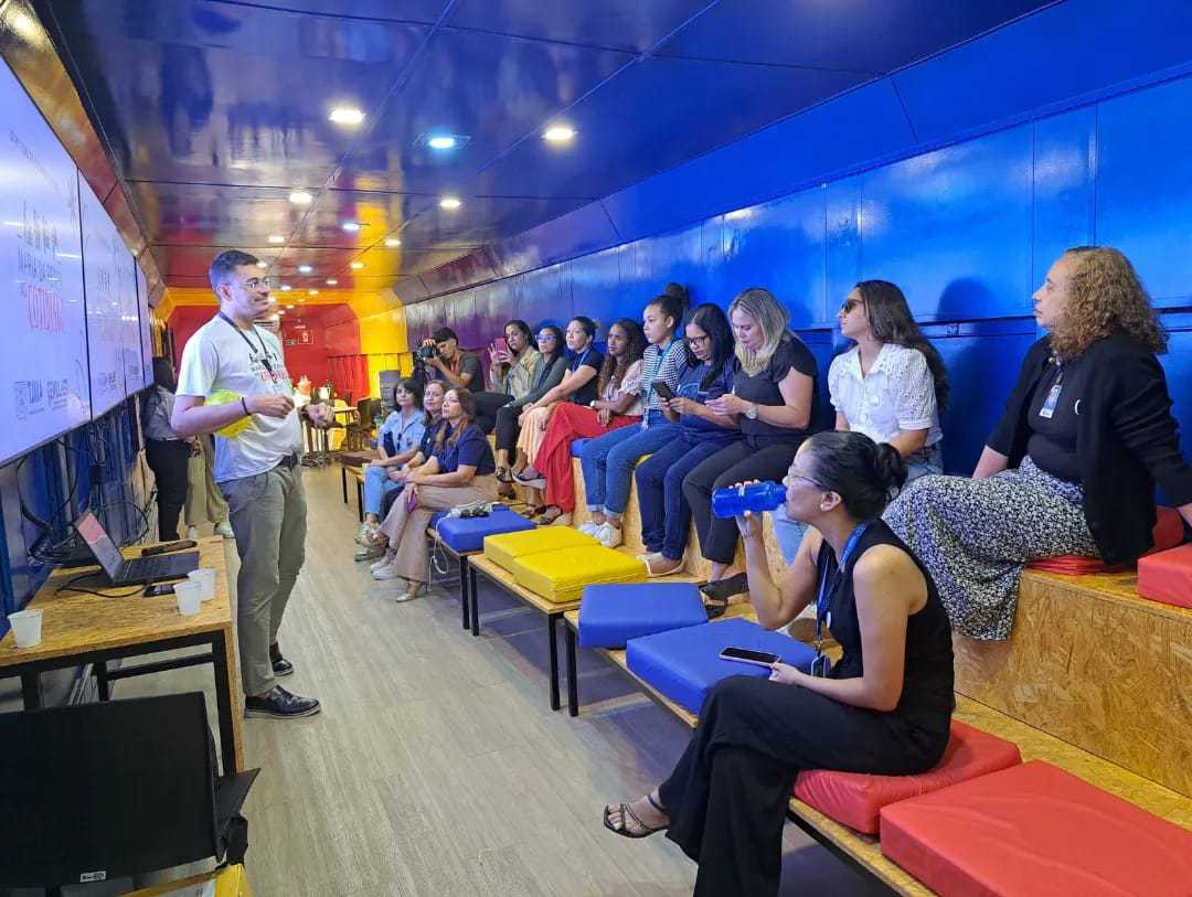 A imagem mostra um grupo de pessoas sentadas em bancos dentro de uma sala com paredes azuis e amarelas, assistindo atentamente a uma apresentação feita por um indivíduo em pé na frente de um quadro branco. O cenário parece ser um ambiente de palestra ou workshop informal, o que pode ser interessante ou relevante para representar uma sessão de aprendizado, reunião comunitária ou evento corporativo.