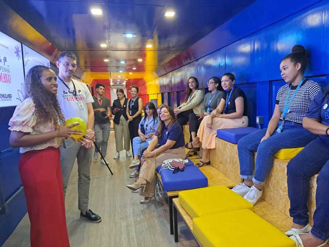 A imagem mostra um grupo de pessoas sentadas em bancos dentro de uma sala com paredes azuis e amarelas, assistindo atentamente a uma apresentação feita por uma mulher em pé na frente de um quadro branco. O cenário parece ser um ambiente de palestra ou workshop informal, o que pode ser interessante ou relevante para representar uma sessão de aprendizado, reunião comunitária ou evento corporativo.