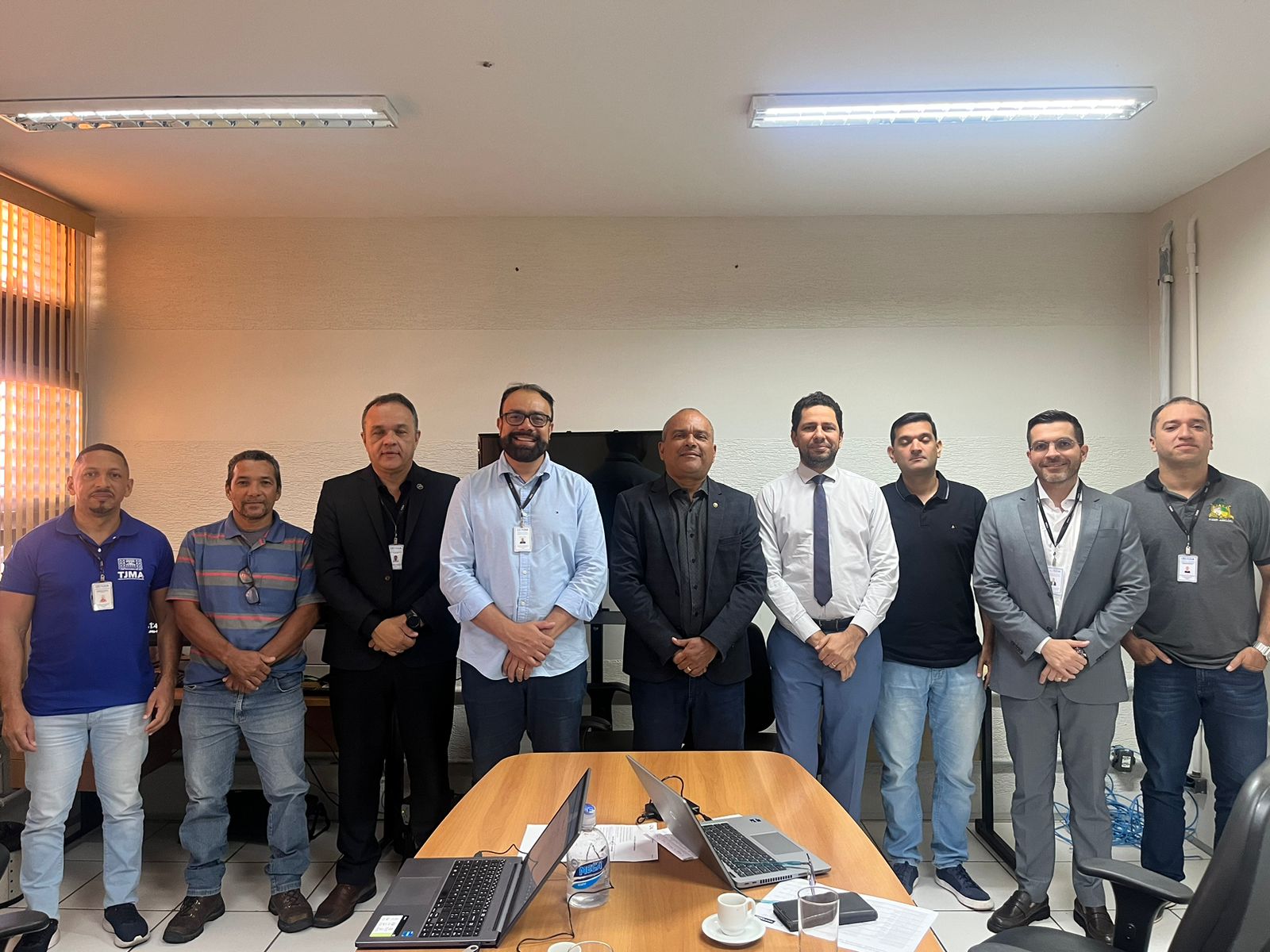 A imagem mostra um grupo de indivíduos em um ambiente de escritório ou sala de conferências. Há nove pessoas no total, todas parecendo ser adultos. Os rostos dos indivíduos foram obscurecidos com blocos coloridos para manter a privacidade. Eles estão em pé na frente de uma mesa com o que parece ser laptops e papéis, sugerindo uma reunião ou encontro relacionado ao trabalho. A sala tem persianas nas janelas, iluminação fluorescente e parece ser um ambiente interno típico para reuniões profissionais