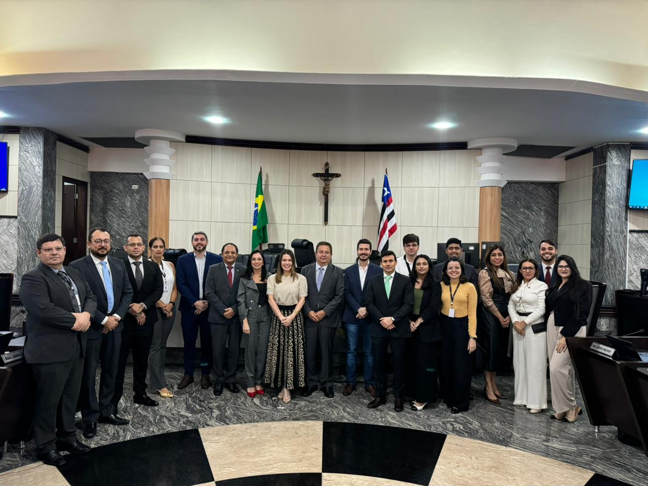 A imagem mostra um grupo de indivíduos em pé em fila, em um ambiente interno, de frente para a câmera. Seus rostos estão obscurecidos por retângulos marrons, provavelmente por motivos de privacidade. Atrás deles, há duas bandeiras exibidas, uma das quais é a bandeira do Brasil. No fundo central, há uma cruz cristã na parede. O cenário parece ser formal e pode ser uma sala de reuniões governamental ou institucional, com base nas bandeiras e na cruz presentes, bem como nas roupas formais dos indivíduos.