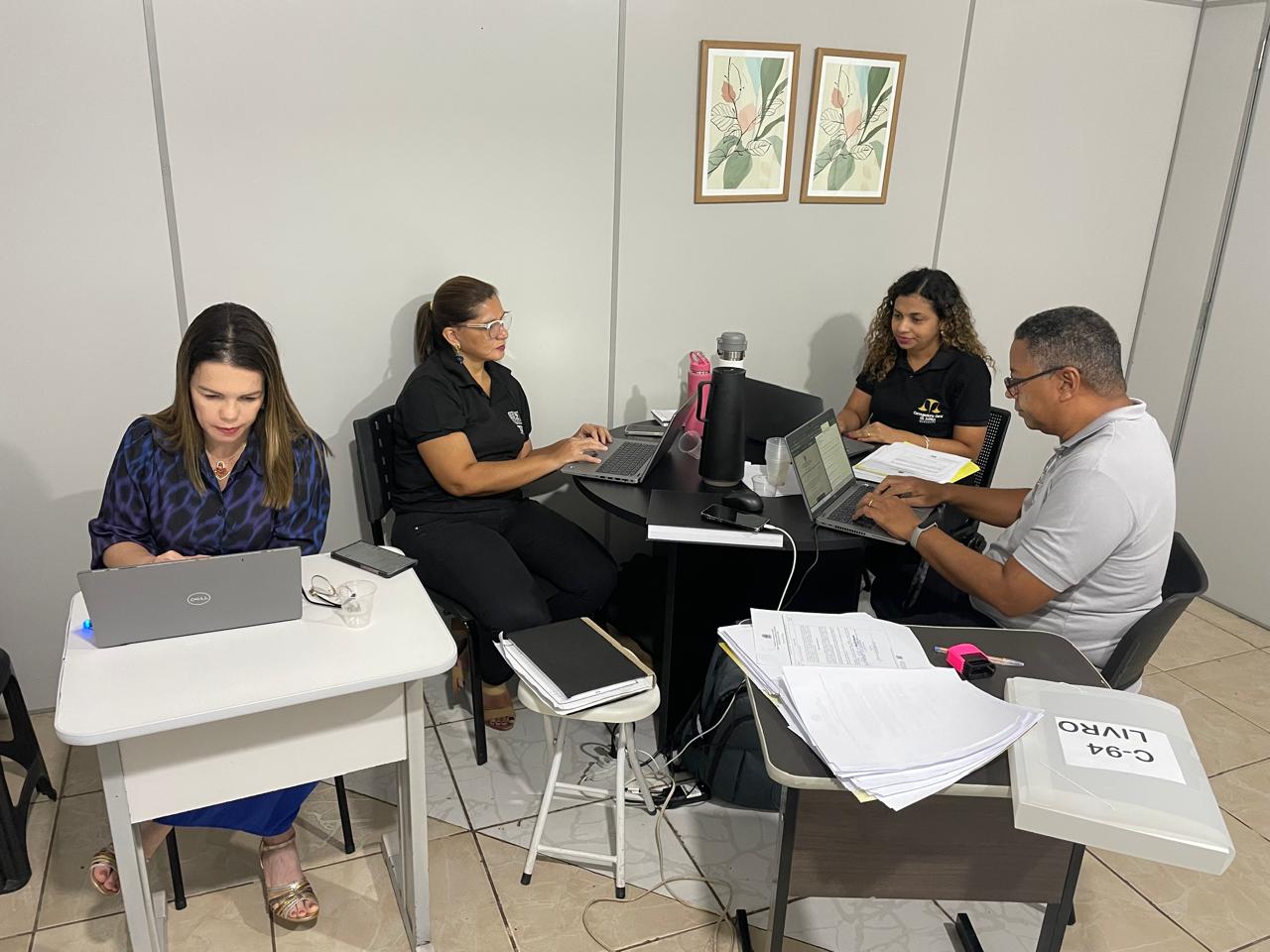 A imagem mostra quatro pessoas sentadas em mesas dispostas em formato de U, cada uma trabalhando em um laptop. Há papéis e cadernos visíveis nas mesas, sugerindo um ambiente de trabalho ou estudo. Os rostos das pessoas foram ocultados com retângulos marrons para preservar a privacidade. Não é possível identificar nenhum problema de matemática ou tarefa visível na imagem.