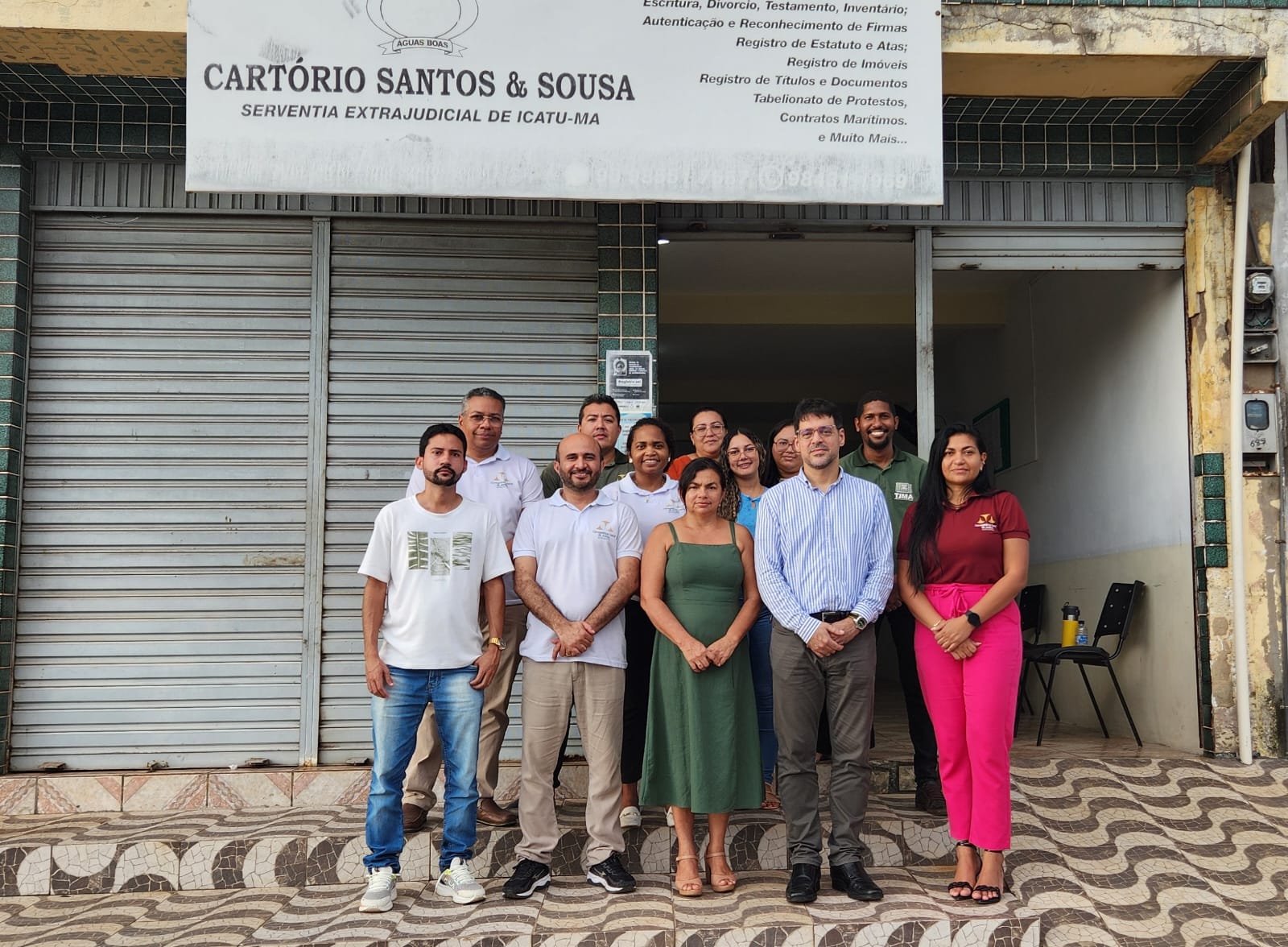 A imagem mostra um grupo de seis pessoas em frente a um prédio com uma placa que diz ¿CARTÓRIO SANTOS & SOUSA SERVENTIA EXTRAJUDICIAL DE CAXIAS-MA¿. As faces das pessoas estão ocultas por retângulos marrons para preservar a privacidade. O prédio parece ser um cartório, conforme indicado pela placa que menciona serviços como ¿Reconhecimento de Firmas, Autenticações, Procurações, Registro de Títulos e Documentos, Registro de Pessoas Jurídicas¿, entre outros.