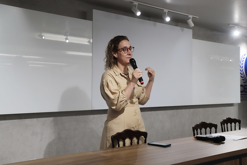 Foto em plano médio de uma mulher de óculos, com cabelos castanhos presos e crespos, falando em um auditório. Ela usa um vestido bege com mangas bufantes. A mulher está em pé, atrás de uma mesa de madeira escura, segurando um microfone com a mão direita e gesticulando com a esquerda, como se estivesse explicando algo. Ela olha para o lado esquerdo, fora do enquadramento. Ao fundo, uma parede de tijolos aparentes cinzas e dois painéis brancos, onde se vê parte de um logo azul de um evento ou instituição.