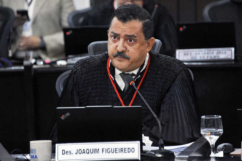 Foto horizontal: Acesso e posse do desembargador Eulálio Figueiredo. A foto mostra um homem sentado, em uma sessão do TJMA. Ele usa uma toga preta com detalhes vermelhos em volta do pescoço e uma camisa branca com gravata preta. Ele tem cabelo escuro penteado para o lado, bigode e está olhando para frente com uma expressão séria. À sua frente, há uma mesa com um microfone, um laptop, um copo com água e um crachá de identificação que diz "Des. JOAQUIM FIGUEIREDO". Ao fundo, outras pessoas e mesas podem ser vistas.