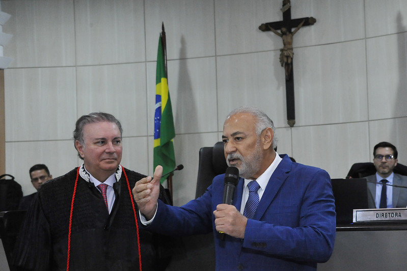 Foto horizontal: Acesso e posse do desembargador Eulálio Figueiredo. A foto mostra uma cena formal, com duas pessoas em primeiro plano. À direita, um homem com barba grisalha e terno azul está falando em um microfone, com a mão esquerda estendida em um gesto. Ele está fazendo um discurso. À esquerda do orador, um homem de cabelo claro, vestido com uma toga preta e detalhes vermelhos, o observa atentamente. Ao fundo, é possível ver uma bandeira do Brasil, um crucifixo na parede, e outras pessoas sentadas. Uma delas está atrás de uma placa que diz "DIRETORA". O ambiente é o plenário do TJMA.
