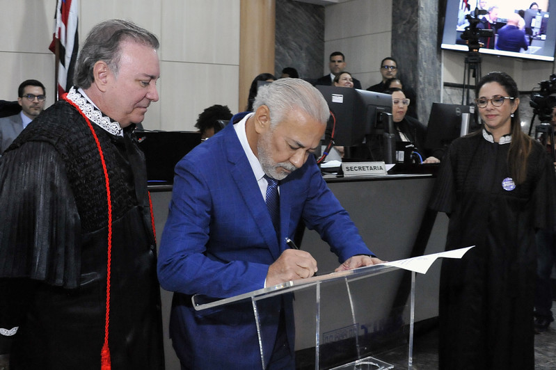 Foto horizontal: Acesso e posse do desembargador Eulálio Figueiredo. A imagem mostra a cerimônia oficial de posse. No centro, um homem com barba grisalha e um terno azul está assinando um documento em uma mesa transparente. À sua esquerda, um homem mais velho, vestido com uma toga preta e detalhes vermelhos, o observa atentamente. À sua direita, uma mulher com óculos e também vestindo uma toga preta, observa a assinatura. Ao fundo, outras pessoas podem ser vistas sentadas e em pé, e há uma placa que diz "Secretaria". O ambiente é o plenário do tribunal.