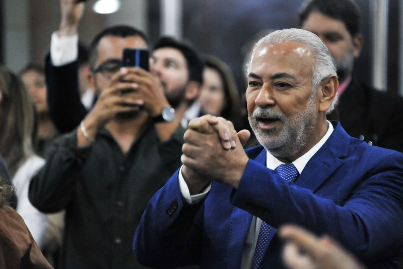 Foto horizontal: Acesso e posse do desembargador Eulálio Figueiredo. A foto mostra um homem com barba grisalha e terno azul, sorrindo e com as mãos juntas na altura do peito, em um gesto que de saudação. Ao fundo, outras pessoas são visíveis, algumas delas segurando celulares para gravar ou tirar fotos.