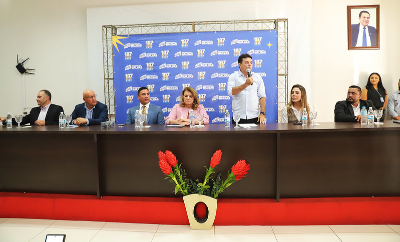 Foto horizontal: TJMA e Prefeitura de Balsas entregam 50 títulos de propriedade. A imagem mostra uma mesa de honra com várias autoridades sentadas, e um homem em pé ao centro, discursando em um microfone. O evento acontece em uma sala com um painel de fundo azul com logotipos da Prefeitura.