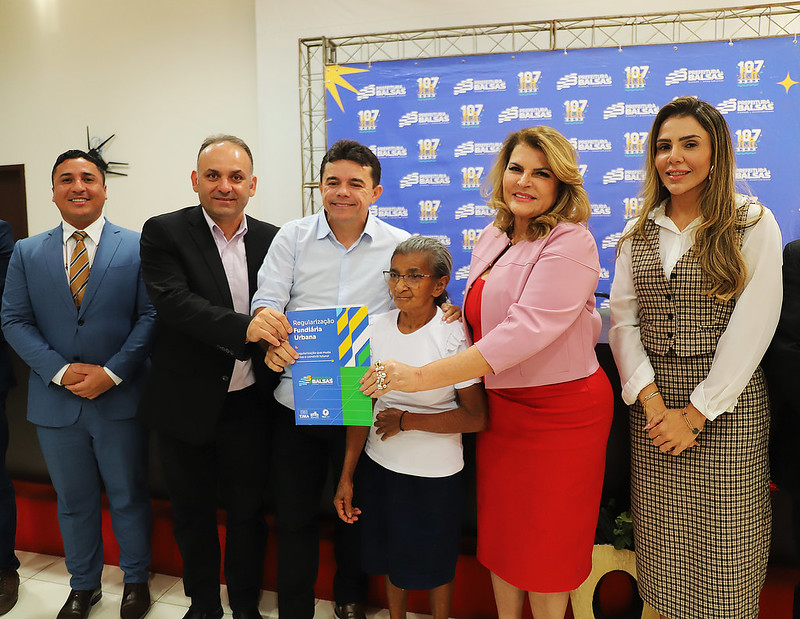 Foto horizontal: TJMA e Prefeitura de Balsas entregam 50 títulos de propriedade. A imagem mostra um grupo de seis pessoas em um evento, com um fundo padronizado com logotipos da Prefeitura.