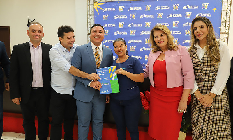 Foto horizontal: TJMA e Prefeitura de Balsas entregam 50 títulos de propriedade. A imagem mostra um grupo de seis pessoas posando para uma foto em um evento. O cenário de fundo tem o logotipo da Prefeitura de Balsas.