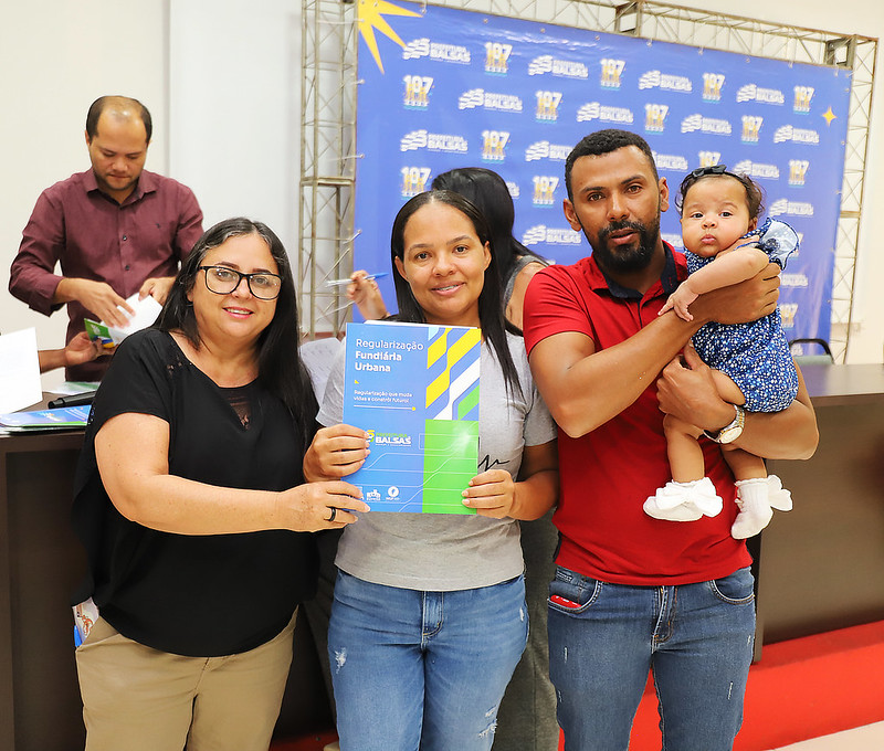 Foto horizontal: TJMA e Prefeitura de Balsas entregam 50 títulos de propriedade. A imagem mostra quatro pessoas em primeiro plano e uma pessoa ao fundo, em um evento semelhante ao da foto anterior, com o mesmo cenário de fundo da Prefeitura de Balsas. Da esquerda para a direita em primeiro plano: Uma mulher de óculos, blusa preta e calça bege, segurando um documento 