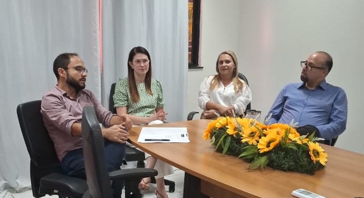 Foto horizontal da Visita da equipe do Prêmio Innovare à equipe da Casa da Criança. A foto mostra quatro pessoas sentadas em uma mesa de reunião. Há dois homens e duas mulheres. Da esquerda para a direita: Um homem com barba e óculos, usando uma camisa social de cor clara com um padrão. Ele está gesticulando com as mãos enquanto fala. Uma mulher de óculos, vestindo uma blusa estampada de tom verde. Outra mulher, usando uma blusa branca. Um homem de óculos, vestindo uma camisa azul, que parece estar ouvindo atentamente. Sobre a mesa, há alguns documentos e um arranjo de flores amarelas e folhagens verdes. O ambiente parece ser uma sala de reunião com cortinas claras ao fundo.