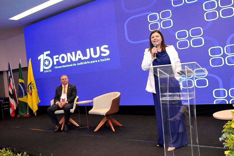 Foto horizontal. Nesta foto, uma mulher, a Conselheira Daiane está em pé à direita, falando em um púlpito transparente com um microfone na mão. Ela veste uma blusa branca sobre um vestido azul escuro e sorri enquanto fala. À esquerda dela, sentado em uma poltrona, está um homem de terno e gravata, com uma credencial pendurada no pescoço. Ao fundo, há um grande painel digital azul com a inscrição "15 ANOS FONAJUS - Fórum Nacional do Judiciário para a Saúde", acompanhado de um logotipo. No canto esquerdo da imagem, há algumas bandeiras, incluindo a do Brasil. A iluminação geral é clara, indicando um evento formal.