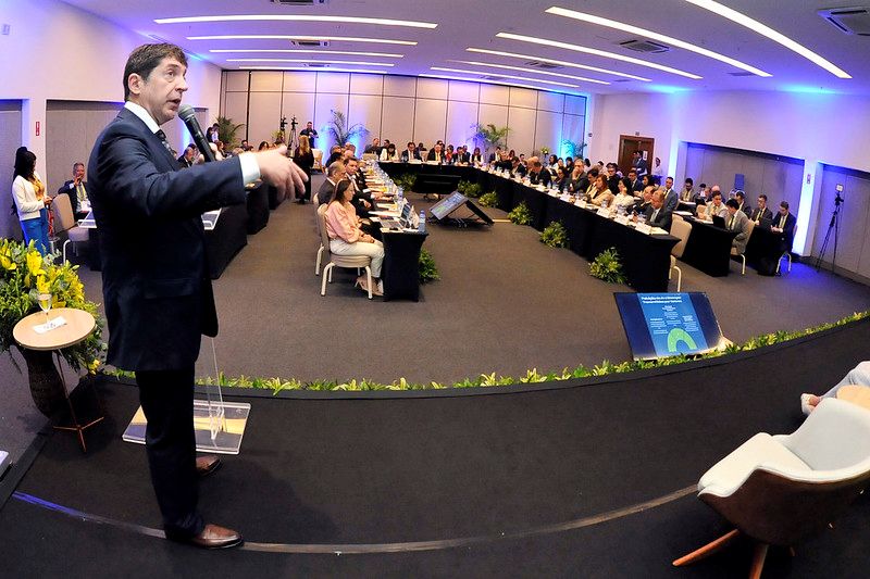 Foto horizontal. Esta foto mostra o "XVI Encontro do Consepre" Nela, é possível ver um homem, o Conselheiro Guilherme Feliciano, em pé na frente de uma sala de conferências, falando para uma plateia de pessoas sentadas em uma mesa longa em formato de "U". A sala é bem iluminada, com luzes azuis e brancas, e há algumas plantas decorativas no chão ao redor da mesa. Há também uma tela de apresentação no lado direito da imagem.