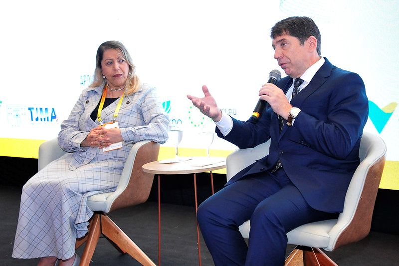 Foto horizontal mostra duas pessoas sentadas em cadeiras de estilo moderno, participando de um evento. O homem à direita, o Conselheiro Guilherme Feliciano, está segurando um microfone e gesticulando com a mão direita, como se estivesse falando. Ele veste um terno azul escuro e uma gravata. Ao seu lado esquerdo, está uma mulher, a presidente do TJBA, sentada com as mãos cruzadas no colo, ouvindo atentamente. Ela veste um conjunto de blazer e calça xadrez em tons claros. Ao fundo, há um painel com logotipos e texto. A iluminação é clara e a cena parece ser de uma apresentação ou painel de discussão.