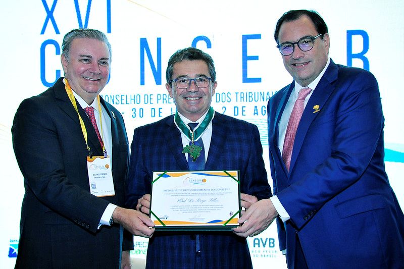 Foto horizontal. Nesta foto, três homens estão em destaque, durante uma cerimônia de entrega de medalha. O homem ao centro está recebendo uma medalha que usa no pescoço e um certificado, entregues pelos outros dois homens, um de cada lado. O homem da esquerda, que veste um terno escuro e uma credencial de evento, está entregando o certificado e olhando para a câmera com um sorriso. O homem do centro, também de terno e óculos, sorri enquanto segura o certificado e exibe a medalha. O homem da direita, que veste um terno azul e uma gravata rosa, também sorri e parece estar entregando a medalha ou auxiliando na entrega do certificado. Ao fundo, há um banner branco com a inscrição 
