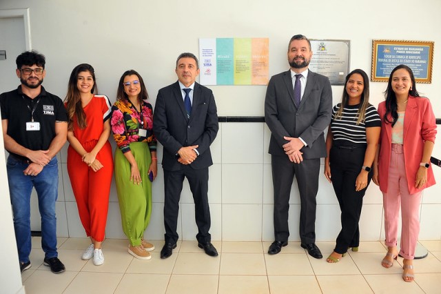 Foto horizontal de servidores, servidoras e de dois magistrados que usam ternos escuros, durante programa Justiça de Proximidade em Buriticupu. Os demais, quatro mulheres e um homem, usam usam roupas de cores e modelos diversos. Entre eles, uma placa com quatro cores. Todos sorriem.