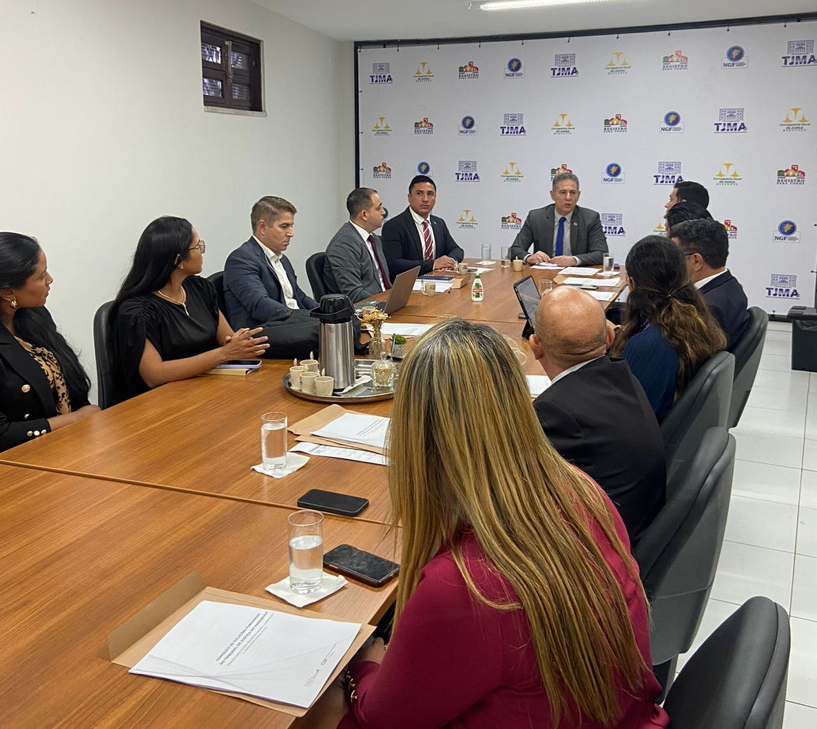 A imagem mostra um grupo de pessoas sentadas ao redor de uma grande mesa de conferência em um ambiente formal de reunião. A sala tem uma aparência profissional, com uma paleta de cores neutras, e há documentos e xícaras na mesa, sugerindo que uma discussão organizada ou negociação está ocorrendo. A parede ao fundo apresenta logotipos, o que pode indicar que a reunião é realizada pela organização representada por esses logotipos.