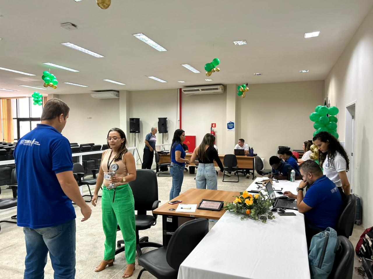 A imagem mostra um ambiente interno que parece ser um escritório ou uma área de atendimento ao cliente. Várias pessoas estão em pé ou sentadas em torno de mesas com computadores. A sala tem paredes brancas e está decorada com balões verdes e amarelos, sugerindo uma ocasião festiva ou comemorativa.