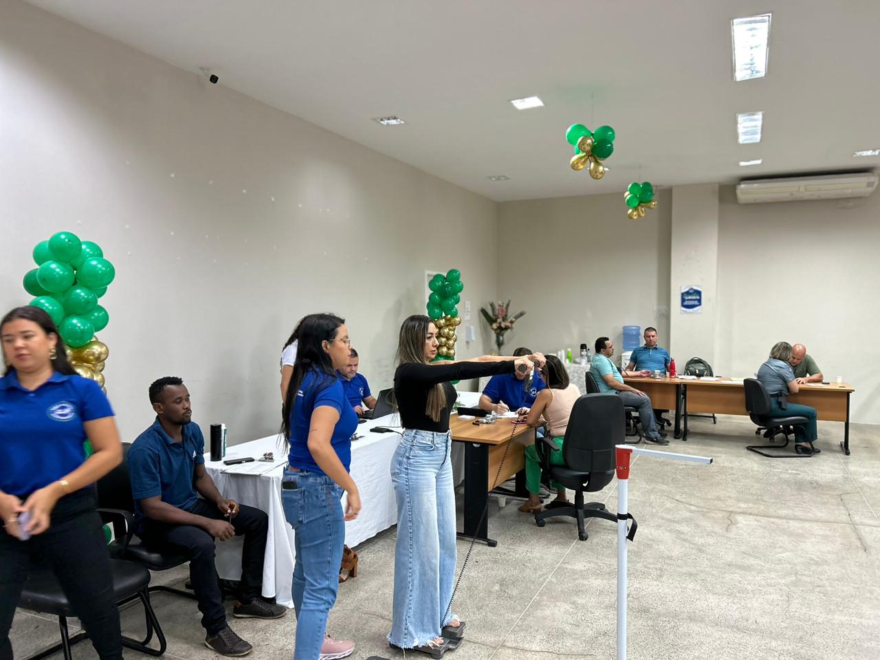 A imagem mostra um ambiente interno que parece ser um escritório ou uma área de atendimento ao cliente. Várias pessoas estão em pé ou sentadas em torno de mesas com computadores. A sala tem paredes brancas e está decorada com balões verdes e amarelos, sugerindo uma ocasião festiva ou comemorativa.