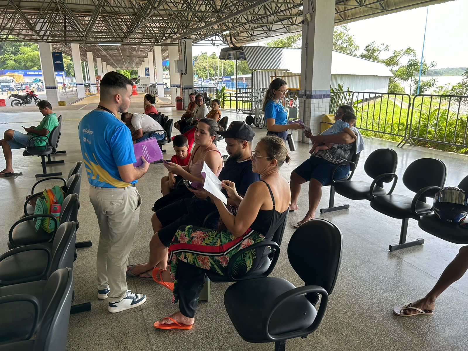A imagem mostra um grupo de pessoas em uma área coberta. Há um grande veículo com o texto ¿CARRETA DA MULHER MARANHENSE¿ na lateral, indicando que é provavelmente uma unidade móvel de serviços de saúde para mulheres no Maranhão. O veículo é rosa e branco, com texto adicional que diz ¿SERVIÇOS GRATUITOS: Mamografia, Preventivo, Exame clínico das mamas.¿ Isso sugere que são oferecidos serviços gratuitos como mamografia, exames preventivos e exames clínicos das mamas. Várias pessoas estão sentadas em mesas, de frente para trabalhadores da saúde ou representantes que estão de pé ou sentados do outro lado. Os trabalhadores da saúde ou representantes parecem estar usando camisas azuis com crachás ou identificações. Os rostos de todas as pessoas foram obscurecidos para privacidade.