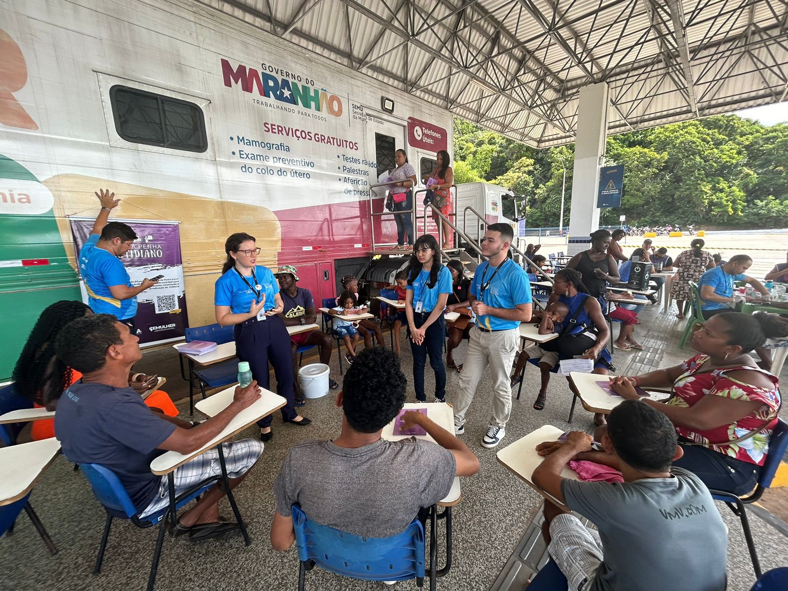 A imagem mostra um grupo de pessoas em uma área coberta. Há um grande veículo com o texto ¿CARRETA DA MULHER MARANHENSE¿ na lateral, indicando que é provavelmente uma unidade móvel de serviços de saúde para mulheres no Maranhão. O veículo é rosa e branco, com texto adicional que diz ¿SERVIÇOS GRATUITOS: Mamografia, Preventivo, Exame clínico das mamas.¿ Isso sugere que são oferecidos serviços gratuitos como mamografia, exames preventivos e exames clínicos das mamas. Várias pessoas estão sentadas em mesas, de frente para trabalhadores da saúde ou representantes que estão de pé ou sentados do outro lado. Os trabalhadores da saúde ou representantes parecem estar usando camisas azuis com crachás ou identificações. Os rostos de todas as pessoas foram obscurecidos para privacidade.