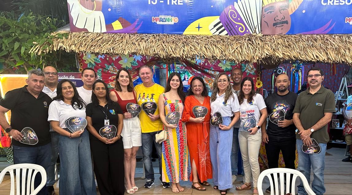 A imagem mostra um grupo de pessoas reunidas para uma foto. Seus rostos estão ocultos por retângulos marrons para proteger a privacidade. Eles estão em um ambiente ao ar livre com decoração tropical, incluindo uma estrutura de telhado de palha e faixas coloridas ao fundo. Alguns indivíduos estão segurando capacetes, sugerindo que isso pode estar relacionado a um evento ou encontro de motociclistas. As faixas têm texto e gráficos, mas o texto não está totalmente visível devido ao ângulo e à distância da foto.