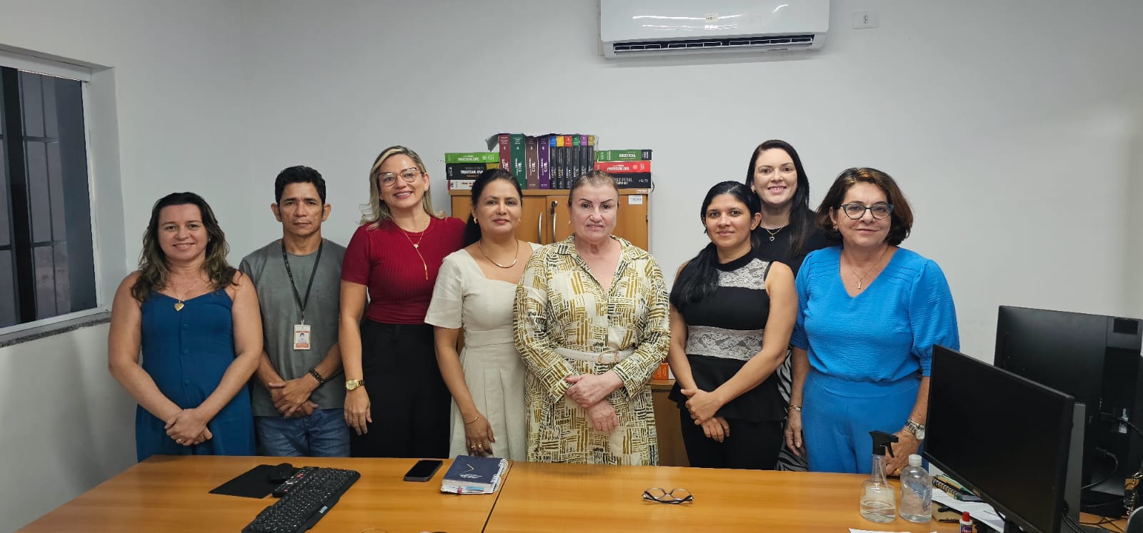 A imagem mostra um grupo de oito pessoas em pé, alinhadas em uma fileira, em um ambiente interno, de frente para a câmera. Parece ser um cenário de escritório, com uma mesa e um computador visíveis à esquerda, e uma estante com vários livros ao fundo. Há um aparelho de ar-condicionado acima deles na parede. A imagem pode ser relevante para representar uma equipe ou membros da equipe em um ambiente de trabalho.