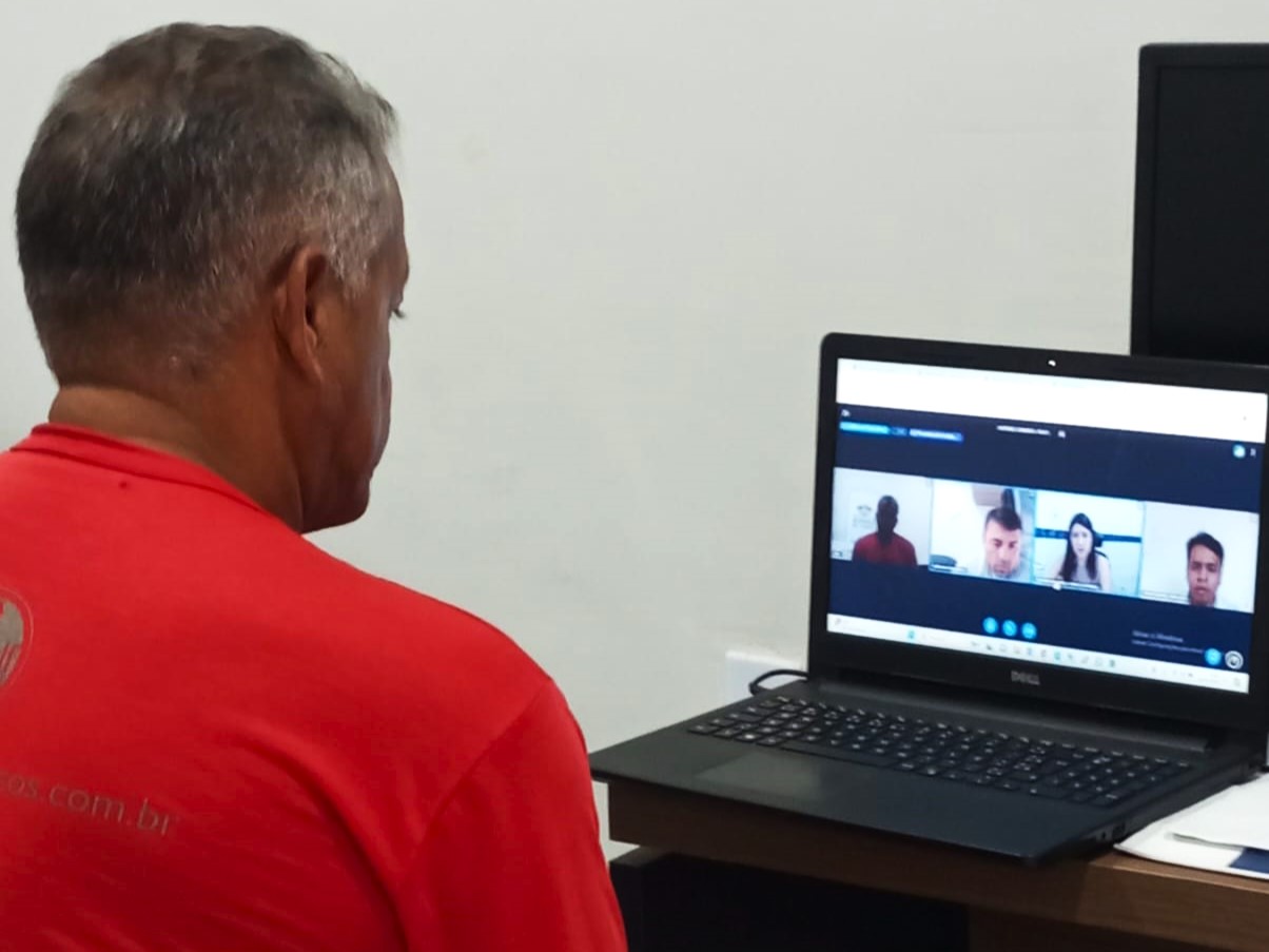 A imagem mostra uma pessoa sentada em uma mesa com um laptop aberto à sua frente. A tela do laptop exibe uma chamada de vídeo em andamento, com outra pessoa visível na tela. À direita do laptop, há outro monitor e uma torre de computador no chão ao lado da mesa. Também há alguns papéis e o que parece ser um caderno ou fichário na mesa ao lado do laptop. O ambiente sugere um escritório ou um espaço de estudo em casa.