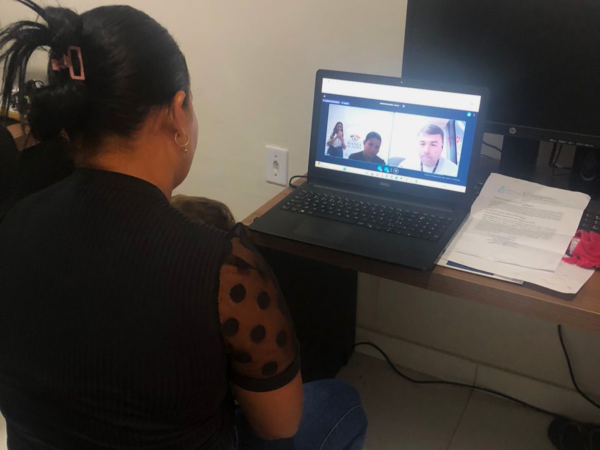 A imagem mostra uma pessoa sentada em uma mesa com um laptop aberto à sua frente. A tela do laptop exibe uma chamada de vídeo em andamento, com outra pessoa visível na tela. À direita do laptop, há outro monitor e uma torre de computador no chão ao lado da mesa. Também há alguns papéis e o que parece ser um caderno ou fichário na mesa ao lado do laptop. O ambiente sugere um escritório ou um espaço de estudo em casa.