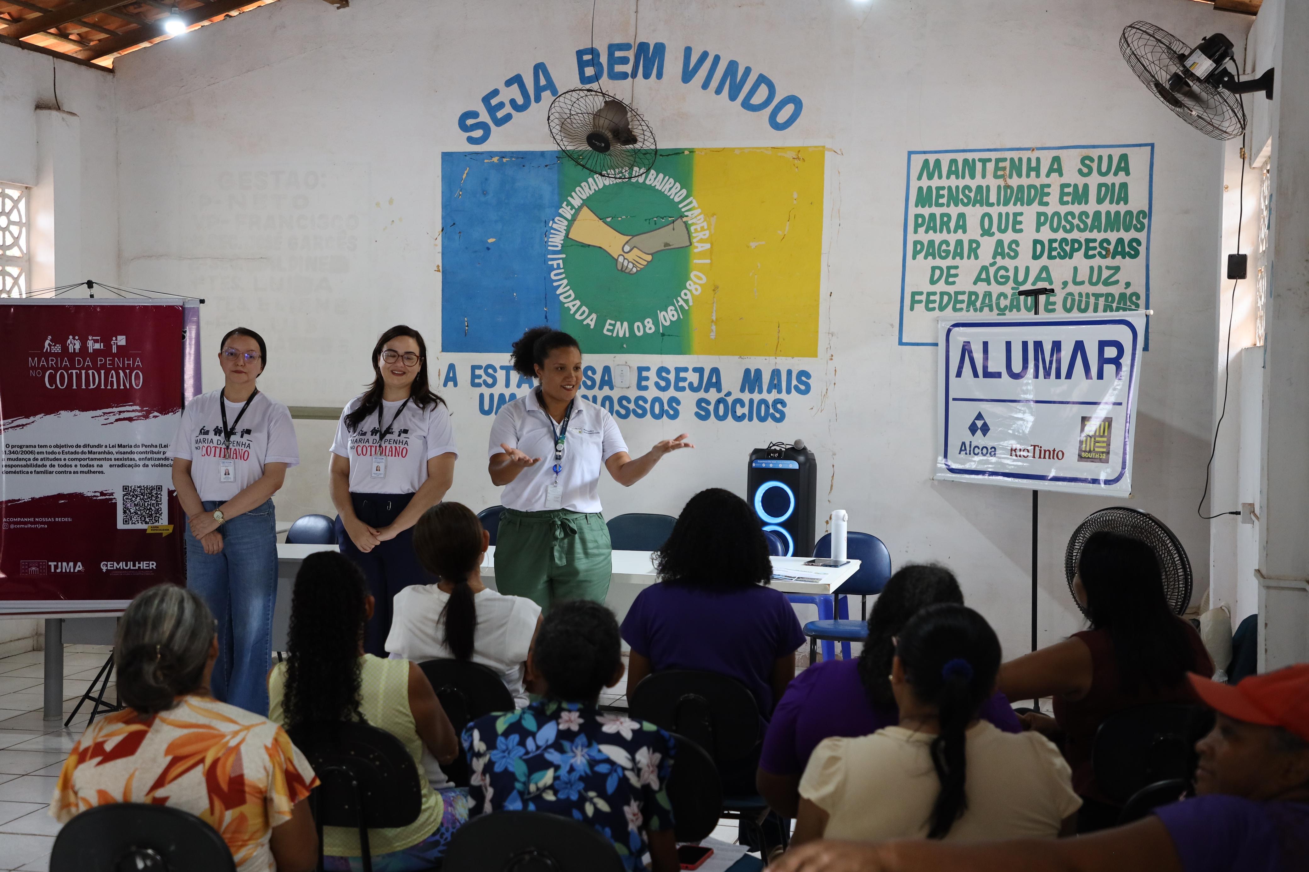 A foto é de dentro de um salão comunitário. Vemos duas mulheres em pé à esquerda, uma usando um óculos e a outra sorrindo, e uma terceira mulher em pé ao centro, de óculos e com um colete branco, que gesticula com as mãos como se estivesse explicando algo. Todas estão voltadas para um público que ocupa várias fileiras de cadeiras azuis, sentadas e de costas para a câmera, prestando atenção. Ao fundo, na parede, há um banner branco com os dizeres em letras azuis "MANTENHA SUA MENSALIDADE EM DIA PARA QUE POSSAMOS PAGAR AS DESPESAS DE ÁGUA, LUZ, FEDERAÇÃO E OUTRAS". Ao lado, há um cartaz da empresa "ALUMAR", com os logotipos da Alcoa e da Rio Tinto. No centro da parede, um banner com a frase "SEJA BEM VINDO" na parte de cima e no centro, um logotipo circular com um aperto de mãos e a data "FUNDADA EM 08/08/1980". Na parede à esquerda, há um cartaz vermelho com a frase "VARA DA MULHER" e a palavra "COTIDIANO" em letras grandes e brancas. No teto, há ventiladores de parede.