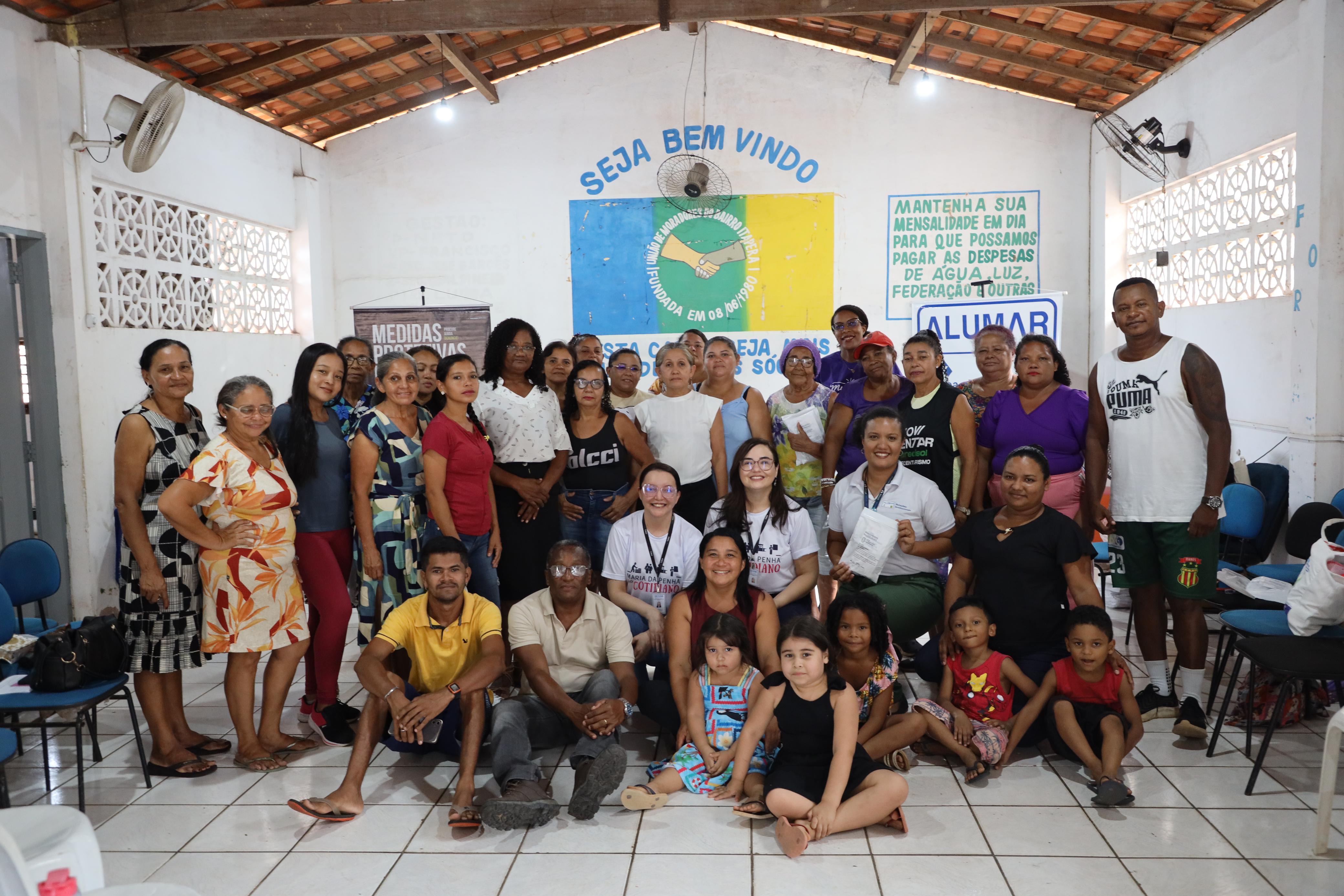 A foto é de um grupo de cerca de trinta pessoas, entre homens, mulheres e crianças, reunidas dentro de um salão. A maioria está em pé, em duas ou três fileiras, e algumas pessoas estão agachadas ou sentadas na frente. O grupo posa para a foto e sorri para a câmera. Ao fundo, as paredes são brancas e há cartazes e banners. Os mesmos banners da primeira imagem estão visíveis: o de "SEJA BEM VINDO" no centro, com o logotipo do aperto de mãos, o cartaz vermelho "VARA DA MULHER", à esquerda, e o cartaz da "ALUMAR", à direita. No teto, é possível ver as vigas de madeira e alguns ventiladores de parede.
