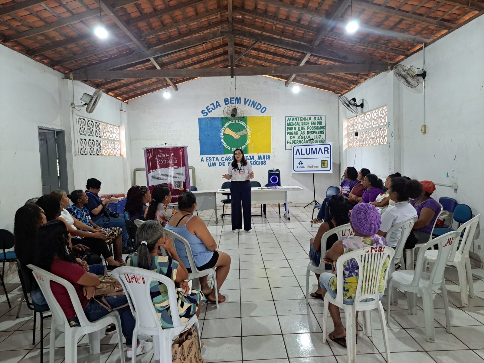A imagem mostra um evento comunitário que acontece em um salão retangular. Ao centro, uma mulher de óculos, com uma blusa branca e calça escura, está em pé, de frente para o público, segurando alguns papéis. Ela parece ser a palestrante. À sua frente, há uma mesa com um notebook preto, um copo de água e uma caixa de som. O público, composto em sua maioria por mulheres, está sentado em várias fileiras de cadeiras brancas de plástico, dispostas nos lados esquerdo e direito do salão. A maioria está prestando atenção na mulher em pé.

Ao fundo da imagem, há uma parede branca com banners. No centro, um banner colorido, dividido em azul, amarelo e verde, traz a inscrição "SEJA BEM VINDO" na parte de cima e um logotipo circular no centro, com um aperto de mãos e a data "FUNDADA EM 08/08/1980". À esquerda, há um banner vermelho com a inscrição "VARA DA MULHER" e a palavra "COTIDIANO" em letras grandes e brancas. À direita, um banner branco com a mensagem "MANTENHA SUA MENSALIDADE EM DIA PARA QUE POSSAMOS PAGAR AS DESPESAS DE ÁGUA, LUZ, FEDERAÇÃO E OUTRAS". Abaixo dele, há um logotipo da empresa "ALUMAR", com os logotipos da Alcoa e da Rio Tinto.

O teto do salão é feito de madeira, com vigas expostas. Há também dois ventiladores de parede, um em cada lado do salão, e luminárias fixadas no teto. As janelas na parte superior da parede, próximas ao teto, têm blocos de cimento vazados, proporcionando ventilação.