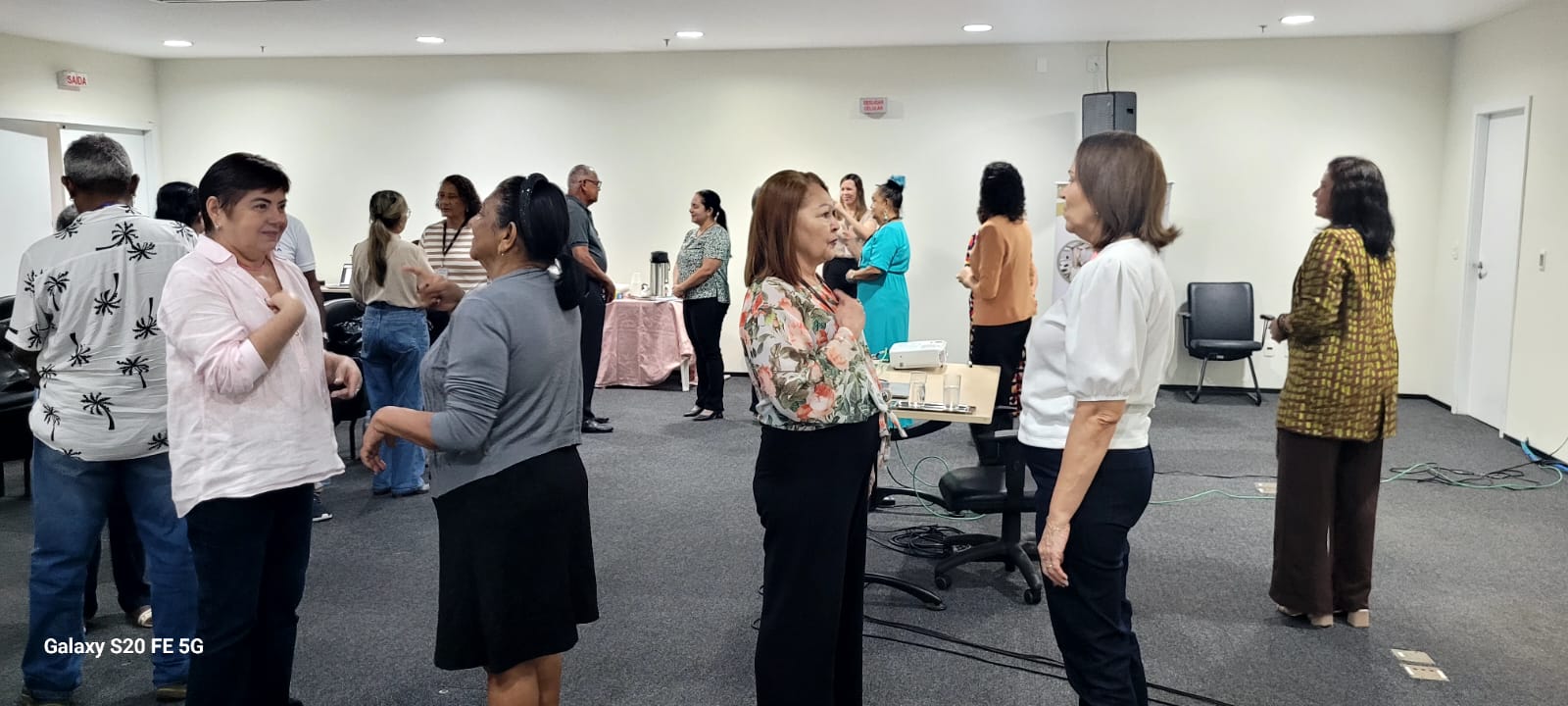 Participantes da oficina interagem em pé, de forma descontraída e sorridente, em duplas ou pequenos grupos. Estão de frente uns para os outros, em dinâmicas corporais. Ao fundo, há uma mesa com café, cadeiras e participantes em movimento.