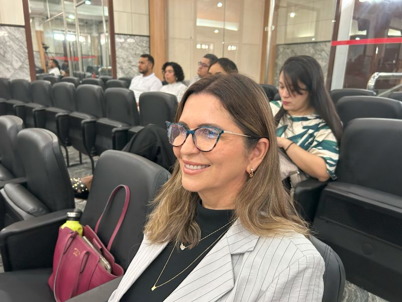 Foto horizntal da juíza Rosânegla Prazeres. A imagem mostra a juíza, sorrindo de lado, sentada em uma das cadeiras de um plenário. Ela usa óculos de armação azul-clara e está com um casaco listrado cinza sobre uma blusa preta, com um colar de pingente dourado. Ao seu lado, numa poltrona, é possível ver uma bolsa magenta. No fundo, há outras pessoas sentadas em fileiras de cadeiras pretas, e o ambiente tem paredes de mármore e iluminação quente.