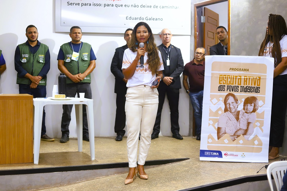Mulher indígena fala ao microfone durante evento do Programa Escuta Ativa dos Povos Indígenas. Ela está ao centro, usa calça clara e camiseta do programa. Ao fundo, outras pessoas acompanham a cerimônia, entre elas integrantes com coletes verdes e servidores do TJMA. Ao lado da oradora, um banner exibe a identidade visual do programa com fotos de uma mulher indígena e uma criança.