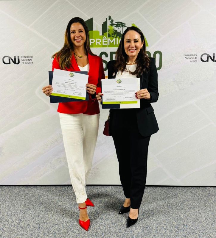 Foto horizontal: TJMA recebe menções honrosas do Prêmio Solo Seguro. A foto mostra duas mulheres em pé, posando com certificados em frente a um painel. O ambiente é de um evento oficial. A mulher à esquerda veste um blazer vermelho sobre uma blusa branca e calças brancas, e segura um certificado na mão. A mulher à direita veste um blazer preto sobre uma blusa branca e calças pretas, e também segura um certificado. Ambas estão sorrindo para a câmera. O painel ao fundo, que serve de pano de fundo para a foto, exibe a logo do "Prêmio Solo Seguro" e os logos do Conselho Nacional de Justiça e da Corregedoria Nacional de Justiça.